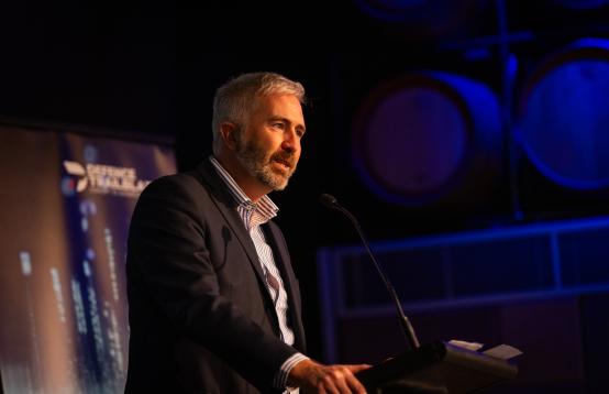 Image of a man with short grey hair and a beard, wearing a dark suit jacket with white business shirt speaking at a podium at a function.