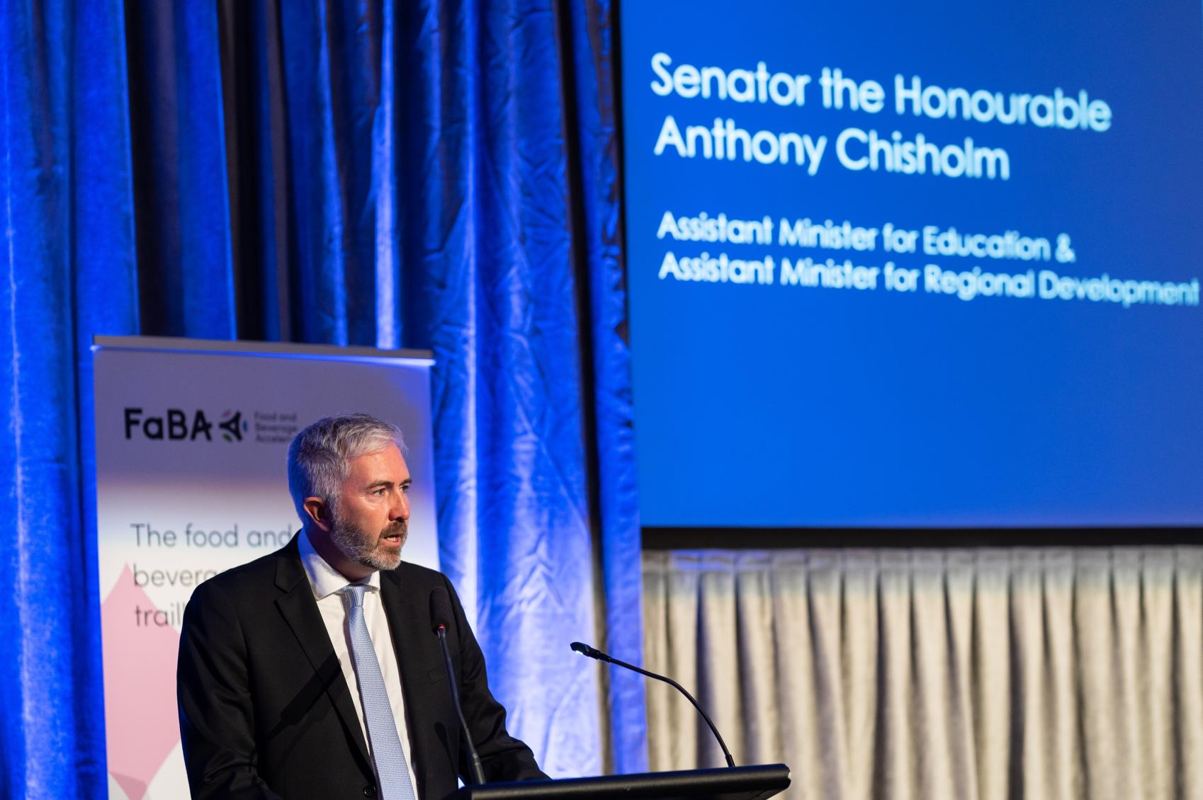 Senator Chisholm wearing a dark suit with a tie standing at a podium presenting to a room
